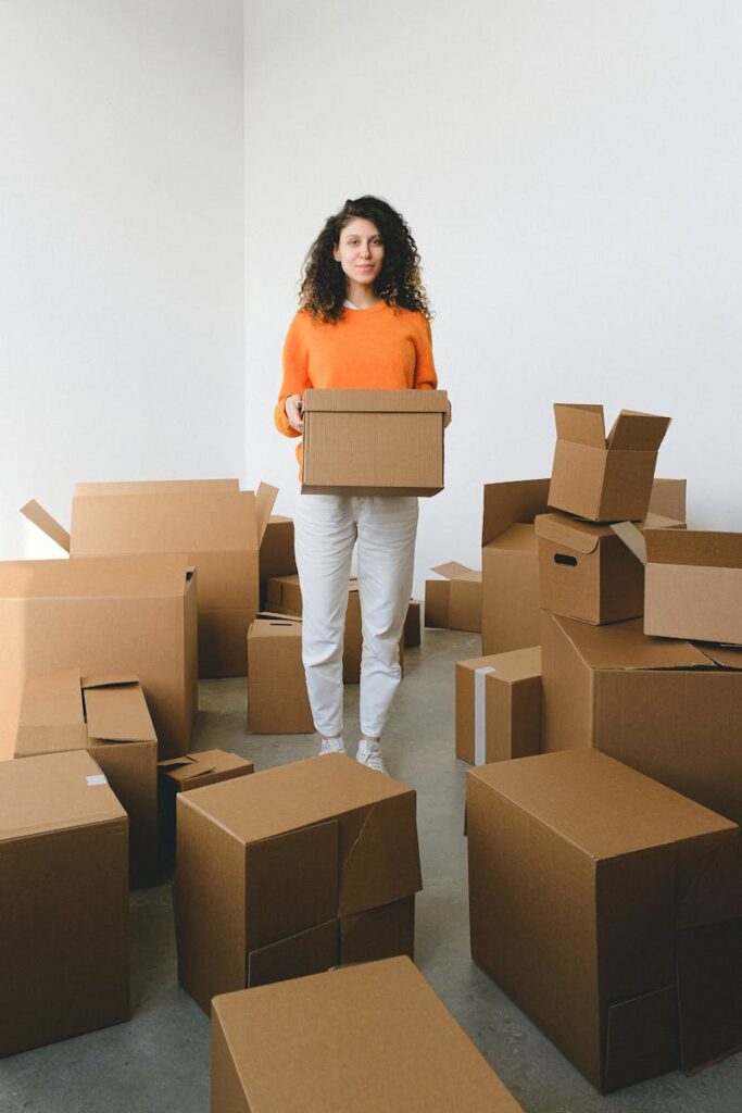 A young woman in a bright orange sweater stands among stacked cardboard boxes, ready to move into her new home.