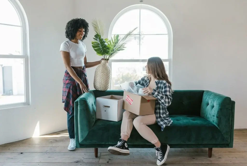 Deux femmes discutent lors d’un déménagement: l’une tient une plante et des boîtes, assise sur un canapé vert, l’autre debout près d’elle. Étapes pour un déménagement réussi.