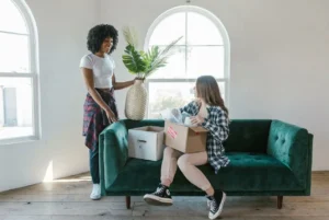 Deux femmes discutent lors d’un déménagement: l’une tient une plante et des boîtes, assise sur un canapé vert, l’autre debout près d’elle. Étapes pour un déménagement réussi.