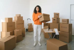 Femme tenant une boîte en carton dans un espace rempli de cartons étiquetés “Books”, “Kitchenware” et “Bedroom” pour un déménagement.
