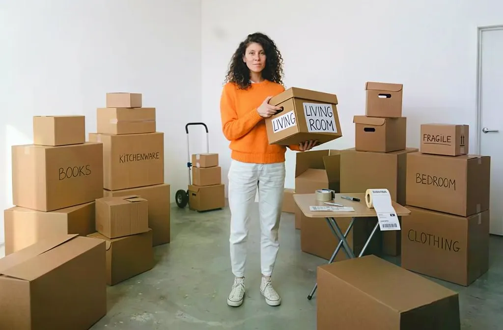 Femme tenant une boîte en carton dans une pièce remplie de cartons étiquetés “Books”, “Living Room” et “Clothing”, prête pour un déménagement. comment emballer les vêtements pour un déménagement
